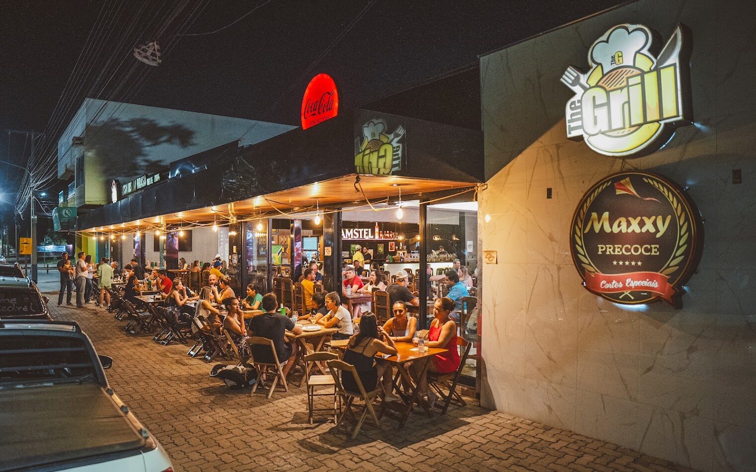 Fachada do The Grill à noite, com mesas na calçada da Av. das Acácias